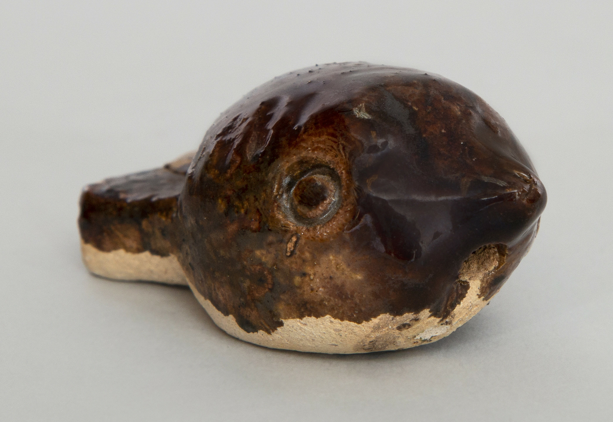 Spaltefløyte av keramikk i form av en liten flat fisk (?). Brun glasur på oversiden, uglasert på undersiden.
Spalte og labium i halsen.
Ingen fingerhull. En tone.