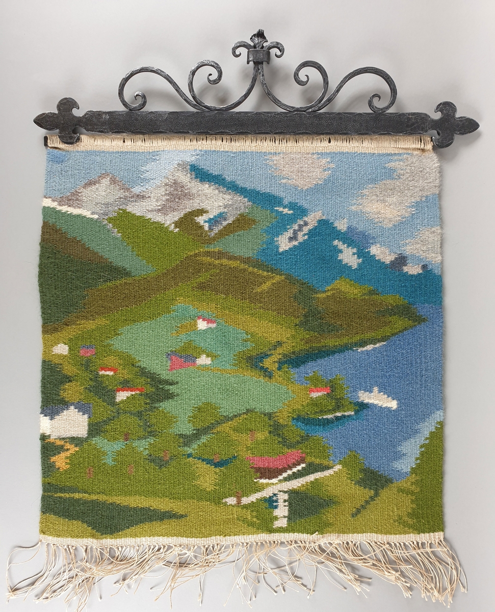 Billedvev med smijernsfeste. Motivet viser kystlandskap med båt, hus, åker, vann, med fjell, himmel og skyer i bakgrunnen