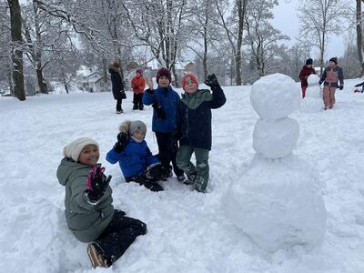 Fire barn er fornøyde etter å ha bygget en snømann med tre kuler oppå hverandre. De jubler, to av dem har armene i været, to sitter på bakken. Det er mye snø og vinter i parken ved Eidsvollsbygningen