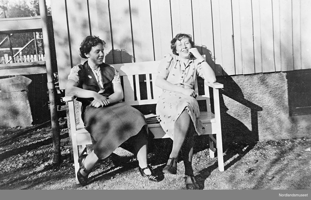 Portrett av Ruth og Randi, familie/venner av familien Ole Vatten. Personene sitter på en benk foran en bolig. Boligen har stående panel. Hagegjerde i venstre billedkant.