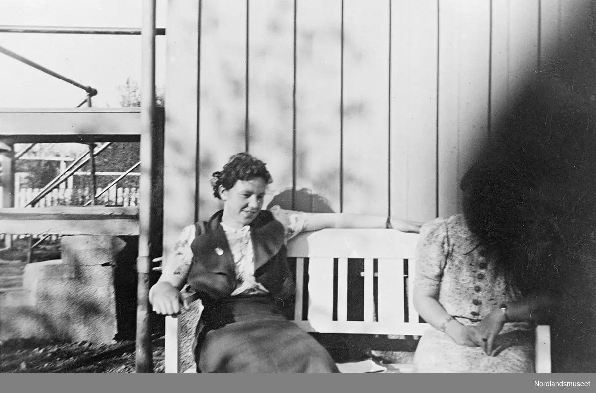 Ruth og Anna Kristine Vatten, datter til Helga og Ole Vatten, på en benk foran en bolig. Boligen har stående panel, dette kan være hjemme hos Anna Kristines foreldre. Hagegjerde i bakgrunnen til venstre.