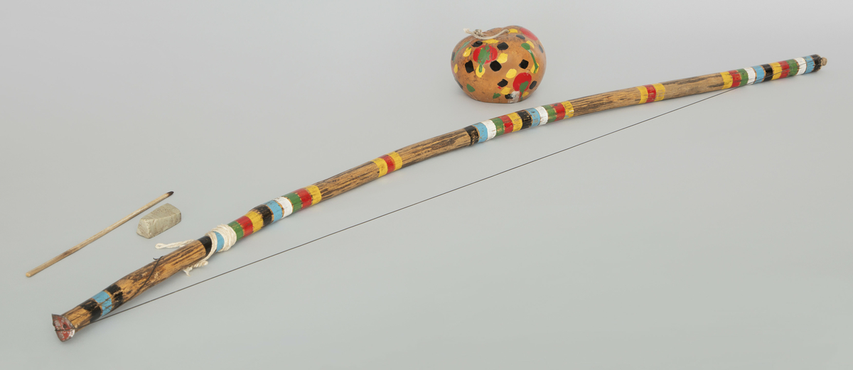 Brasiliansk musikkbue av tre med resonator av kalebass og streng. Medfølgende stein og stikke brukes ved spill på buen. Buen og resonatoren er dekorert med striper og prikker i svart, blått, hvitt, grønt, rødt og gult. 