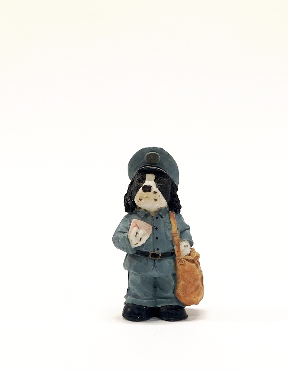 Figurin förställande en hund i uniform med mössa och brevbärarväska. Hunden håller ett brev.