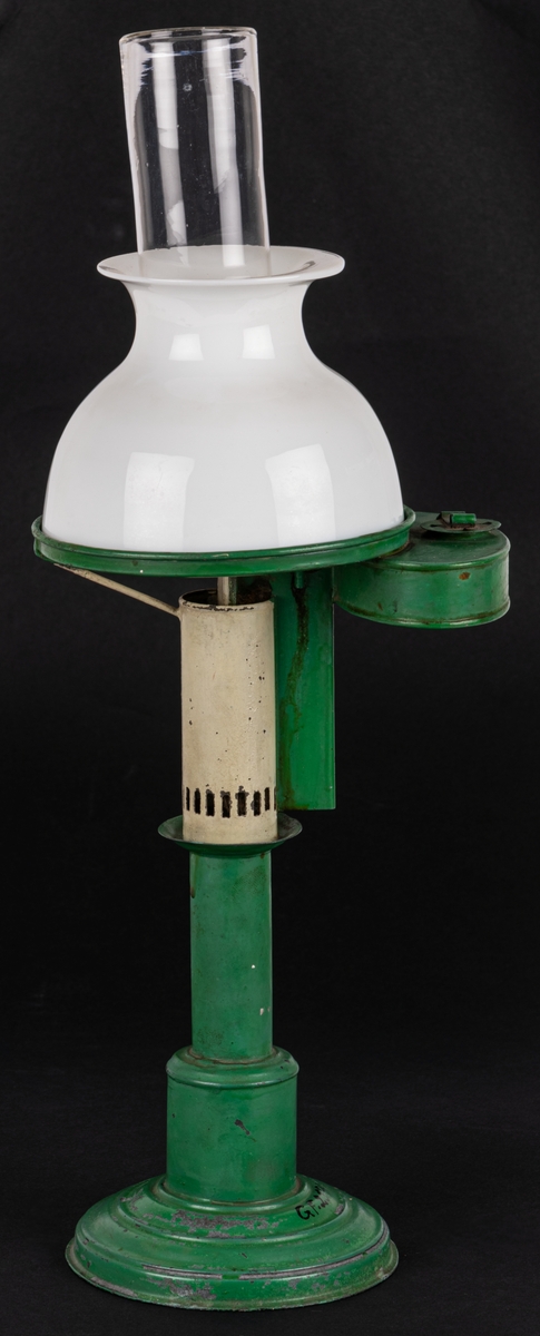 Bordslampa, liten, grön och vit.
Grönmålad plåt, på rund fot med njurformig behållare vid sidan i höjd med den lilla vita porslinskupan. Rakt, cylindriskt brännarglas.