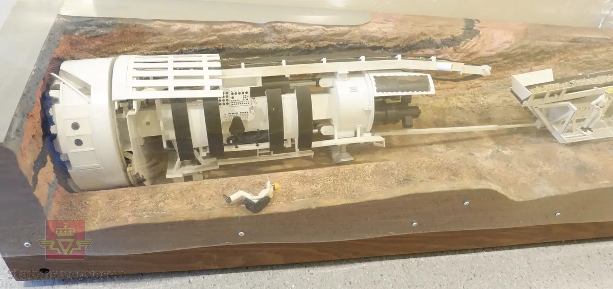 Miniatyrmodell av Atlas Copco fullprofil, tunnelbormaskin. Maskinen er plassert på en sokkel av treverk inni en bue av pleksiglass. Modellen viser tunnelbormaskinen i arbeid med to arbeidere i en boringsprosess. En skiltplate er plassert på sokkelen, og har innskriften SCALE MODELS, INC CLEVELAND, OHIO.