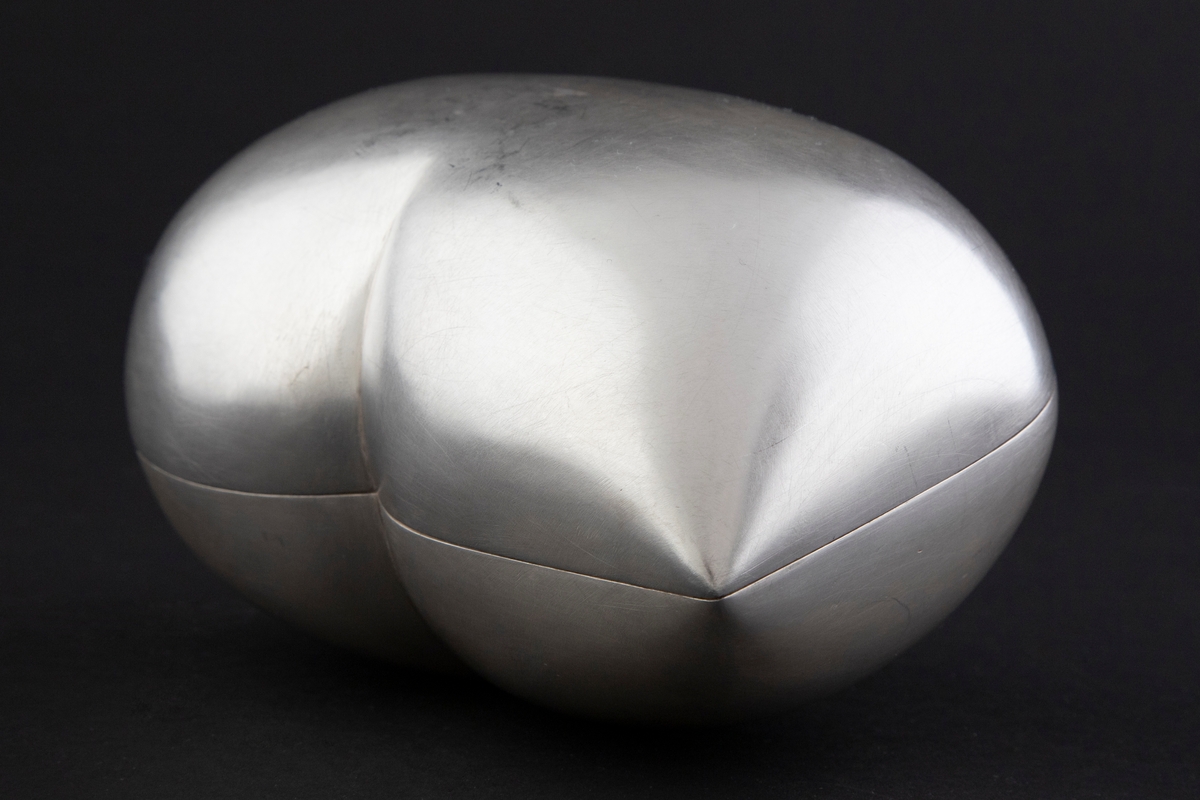 Bonnbonière i sølv. Oval, avrundet og voluminøs skulpturell form, som munner ut i en spiss på den ene siden. Lokkets og bunnens former speiler hverandre. Overflaten er matt, mens på innsiden har overflaten en hamret struktur.