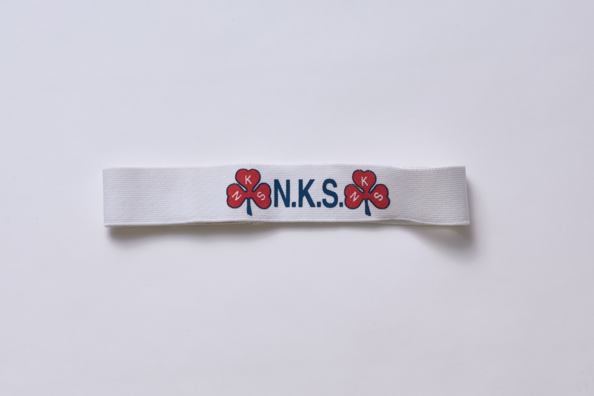 Tøyelig svettebånd med Norske Kvinners Sanitetsforenings logo i form av et rødt trekløver. Glatt utside, mens innsiden er flosset. 