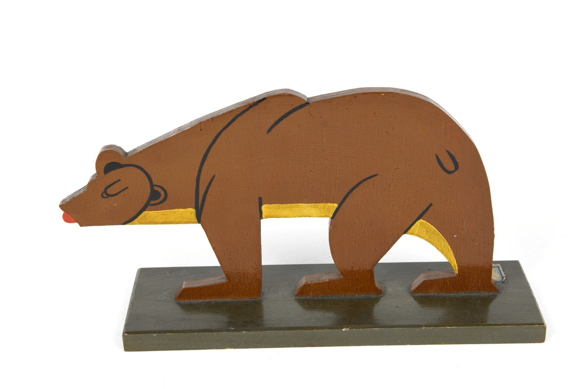 En bjørnefigur laget av  en tynn treplate. Bjørnen er brunmalt med gul/gyllen buk. Ører, øyne, hode, hale og for-/bakbein er markert med svart. Tungen stikker ut og er rødmalt. Den er montert stående/gående på en firkantet svart tresokkel.