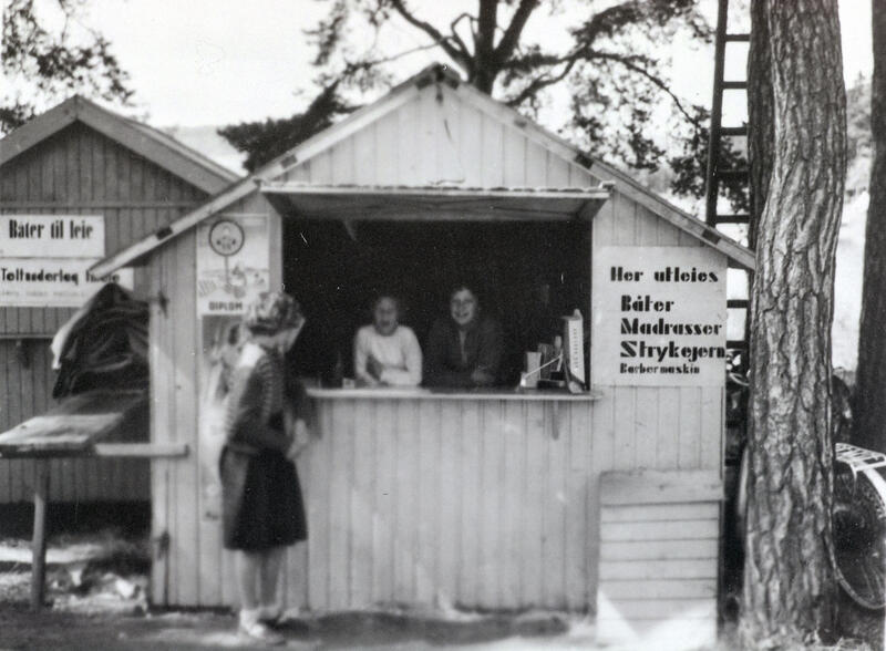 Kioskbod med to personer i luka og en kunde utenfor. Plakat til høyre for luka annonserer med utleie av båter, madrasser, strykejern og barbermaskiner.