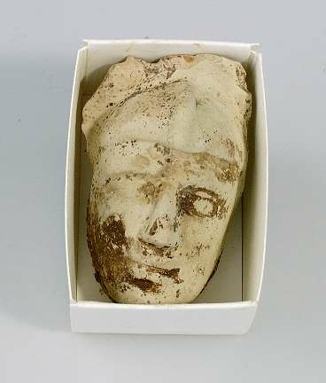 Keramikfragment i form av ett ansikte i ljust lergods. Har suttit på något föremål.
