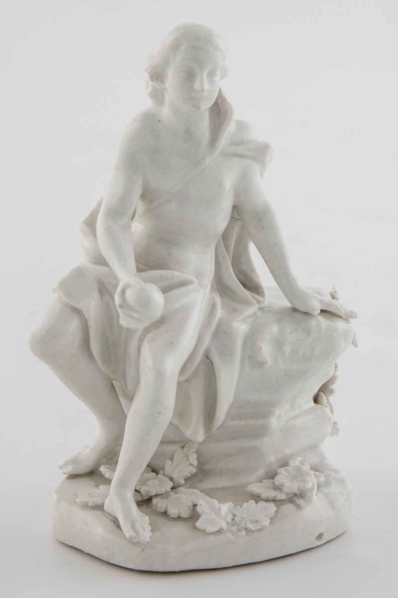 Hvitglasert, umalt porselensfigur med en ung mann sittende på en klippe. Figuren holder et eple i høyre hånd og identifiseres som den trojanske prinsen Paris fra gresk mytologi. Sokkelen er dekorert med blomster og blader.