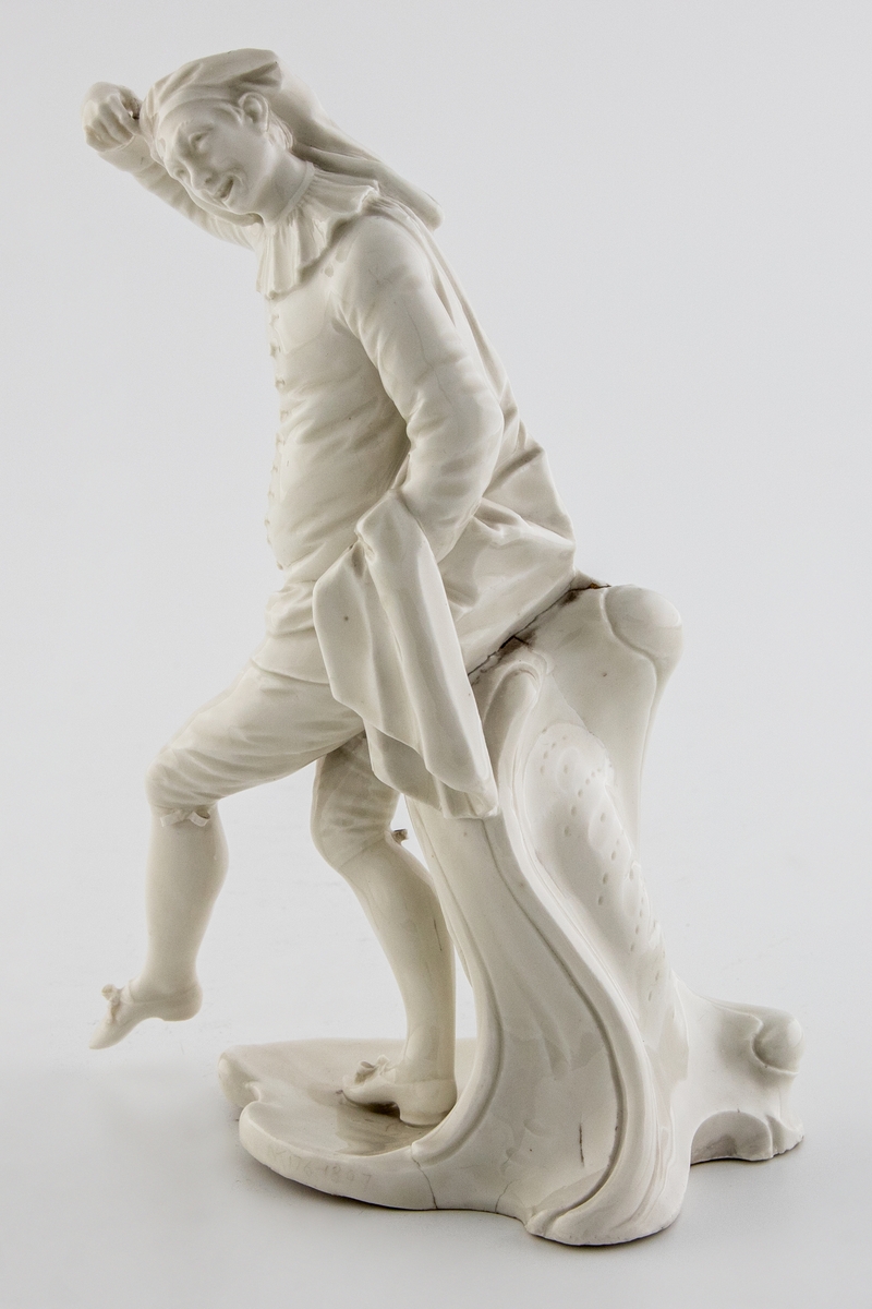 Hvitglasert, umalt porselensfigur av en dansende mann i 1700-talls klær som støttes av et postament med rokokko-trekk. Med høyre hånd tar han på sin lue og med venstre hånd holder han kappen åpen. Figuren beskrives både som harlekin, og identifiseres som scaramuzzio (også: scaramuccia, scaramouche eller scaramuz), en karakter fra commedia dell´arte som oftest blir fremstilt i svart spansk drakt.