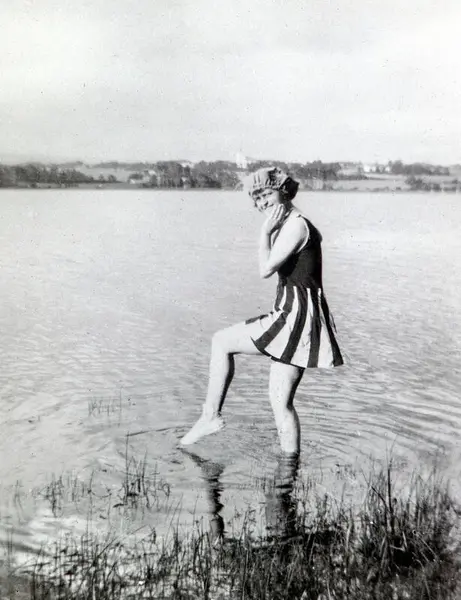 Svart-hvitt foto av ung kvinne iført badedrakt med skjørt, som putter tåa i vannet.