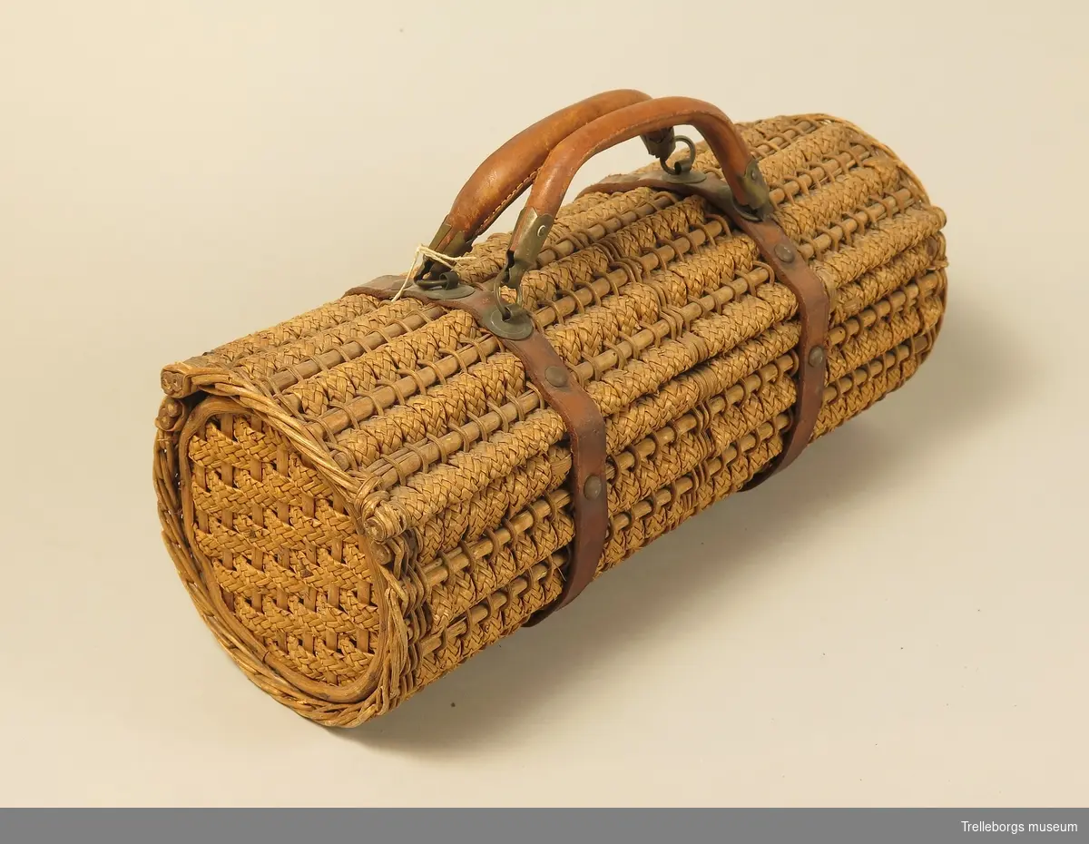 Väska med läderremmar och läderhandtag. Väskans sidostycke utgöres av två st cirkelrunda träplattor. Väskans stomme utgöres av smala pinnar kring vilka det trätts band (0,8 cm) av flätade strån.