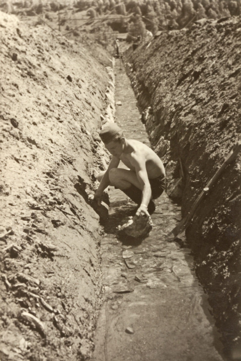 En mann arbeider på en vanningskanal. Kanalen i senter av bildet, 1 mann i bar overkropp med  militærlue arbeider på i bunnen av kanalen. Skogkledd åsside.