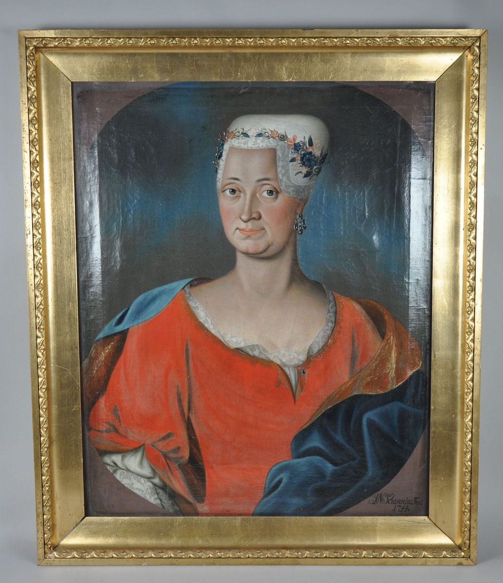 Stående portrettmaleri. Frontalportrett av dame i rød kjole og hvit hodeplagg med blomsterdekor. Damen har øredobb. 
Maleriet har gullbelagt, profilert ramme av tre.