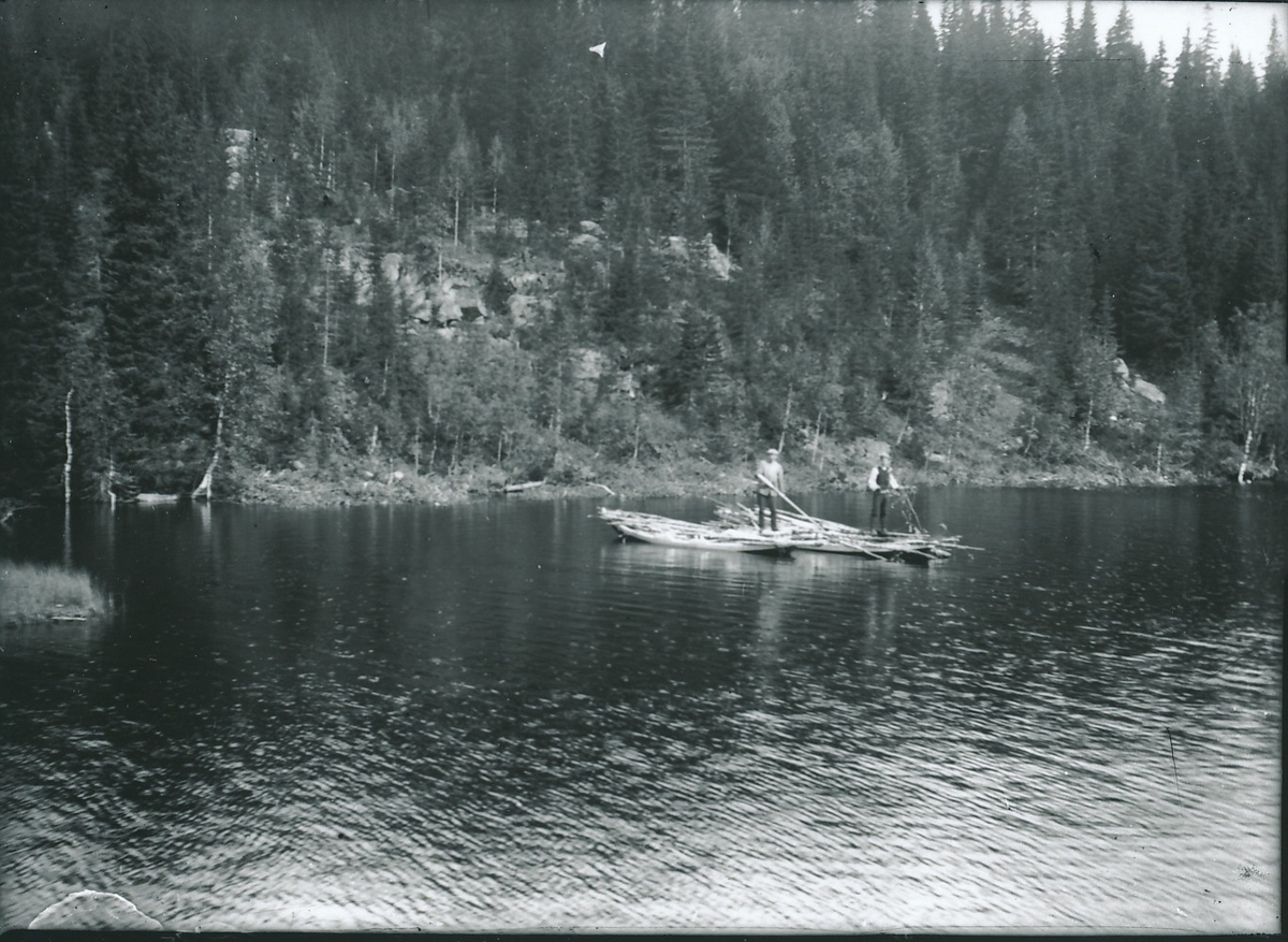 To menn står i hver sin båt (eike) ute på innsjø. Begge båtene er lastet med trestokker. I bakgrunnen er en fjellside med skog.