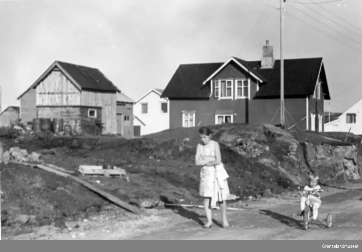 Bugøynes 17.08.1970.
Elsa og Kjell-Aksel Haldorsen på veien nedenfor nyskolen.
I bakgrunnen Aina Dørmænens hus og fjøs sett fra sør-vest.