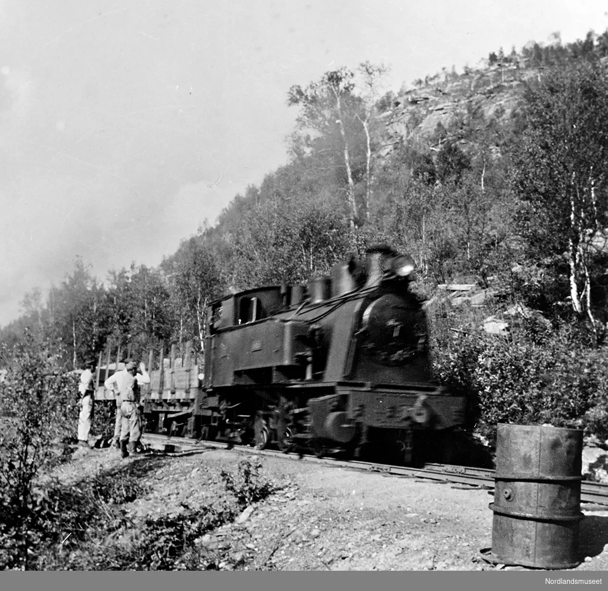 Damplokomotivet Saulo, fra Sulitjelmabanen, tog i fart. Toget passerer noen linjearbeidere. I nedre høyre hjørne står et oljefat.