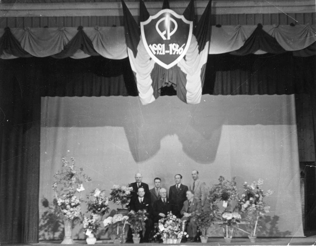 Bild på sju män som står eller sitter i två rader på en scen. I taket flaggor och ett emblem där det står FP 1921-1946. Förmodligen från Linköping Folkets Parks 25-årsjubileum.