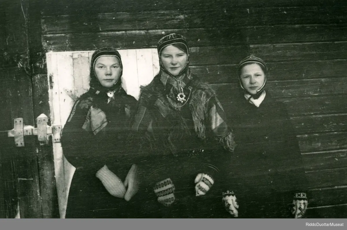 Golbma sámi nieidda olggobealde viesu.
Tre jenter står ved veggen.