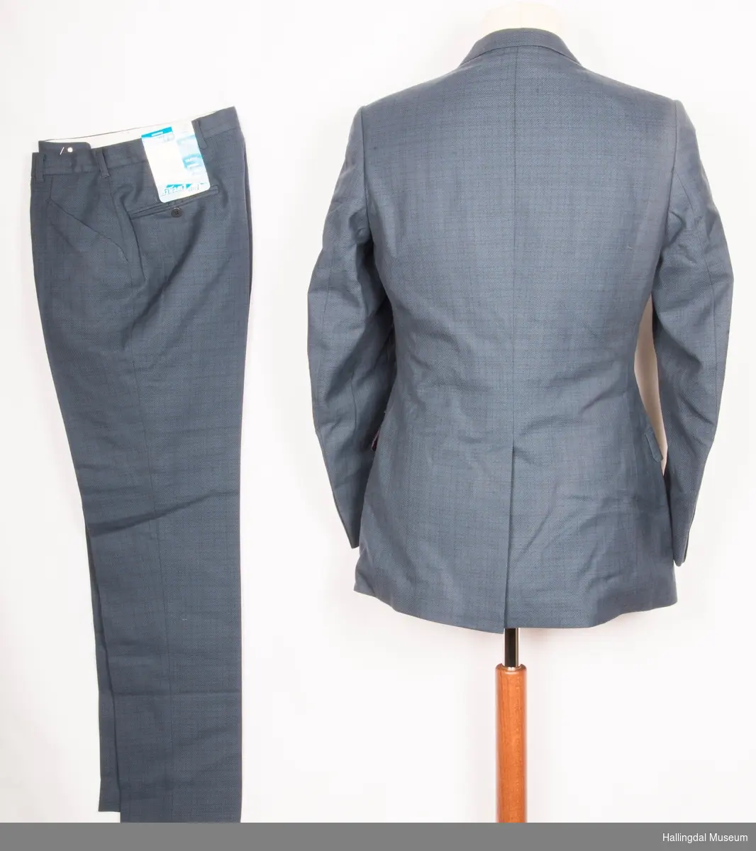 Grå herredress med jakke og bukse fra K. Grudes konfeksjonsfabrikk AS.  Dobbeltspent dressjakke med 3 knapper og knapphull på hver side.
