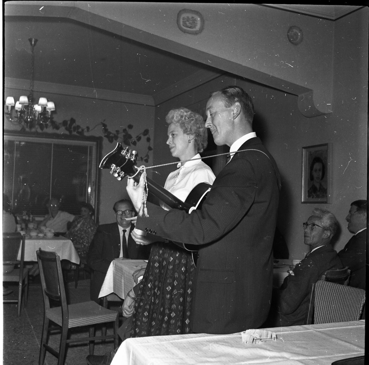 En man och en kvinna står framför en sittandes publik. Mannen spelar gitarr och kvinnan sjunger.