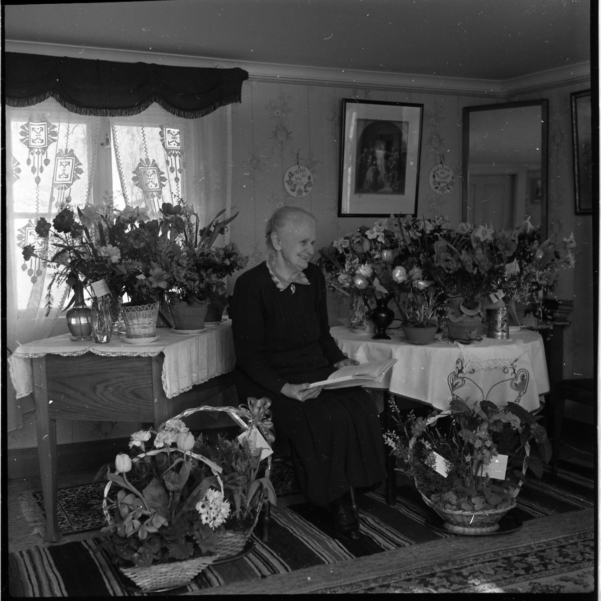 En kvinna sitter omgiven av blombuketter och blomsterarrangemang. Hon håller i en bok. Hon bär en svart klänning med spetskrage. hon tittar på en punkt bortanför kameran och skrattar.