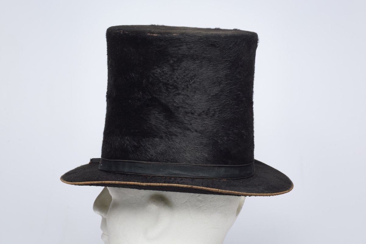 Flosshatt (a) og hatteeske (b). Flosshatten er svart og har et pyntebånd med sløyfe. Innvendig er den foret med en bred lærkant og lyst stoff. Hatteesken er av papp, mørk brun og av samme fasong som hatten. Inne i hatten er et produsentstempel med en krone over navnet.