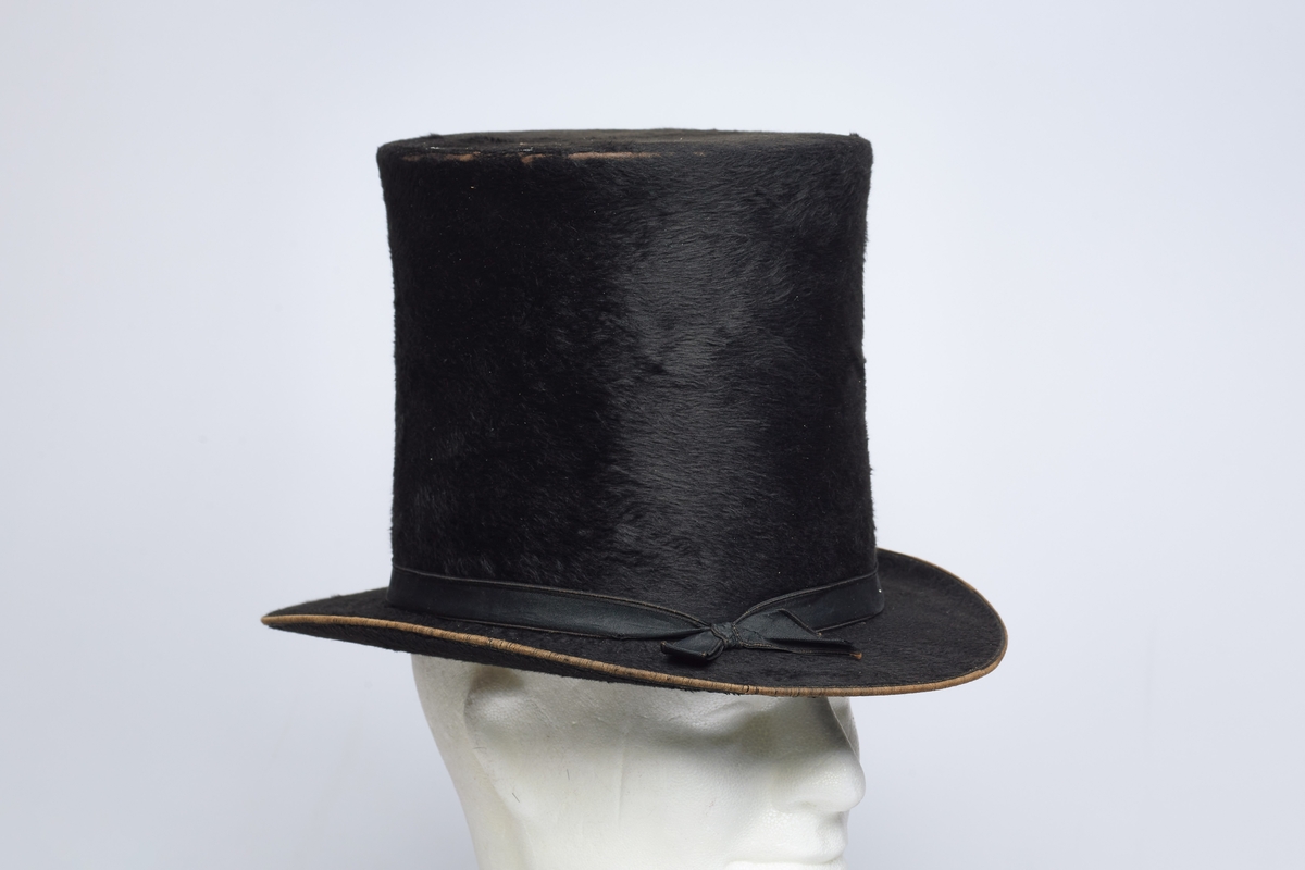 Flosshatt (a) og hatteeske (b). Flosshatten er svart og har et pyntebånd med sløyfe. Innvendig er den foret med en bred lærkant og lyst stoff. Hatteesken er av papp, mørk brun og av samme fasong som hatten. Inne i hatten er et produsentstempel med en krone over navnet.