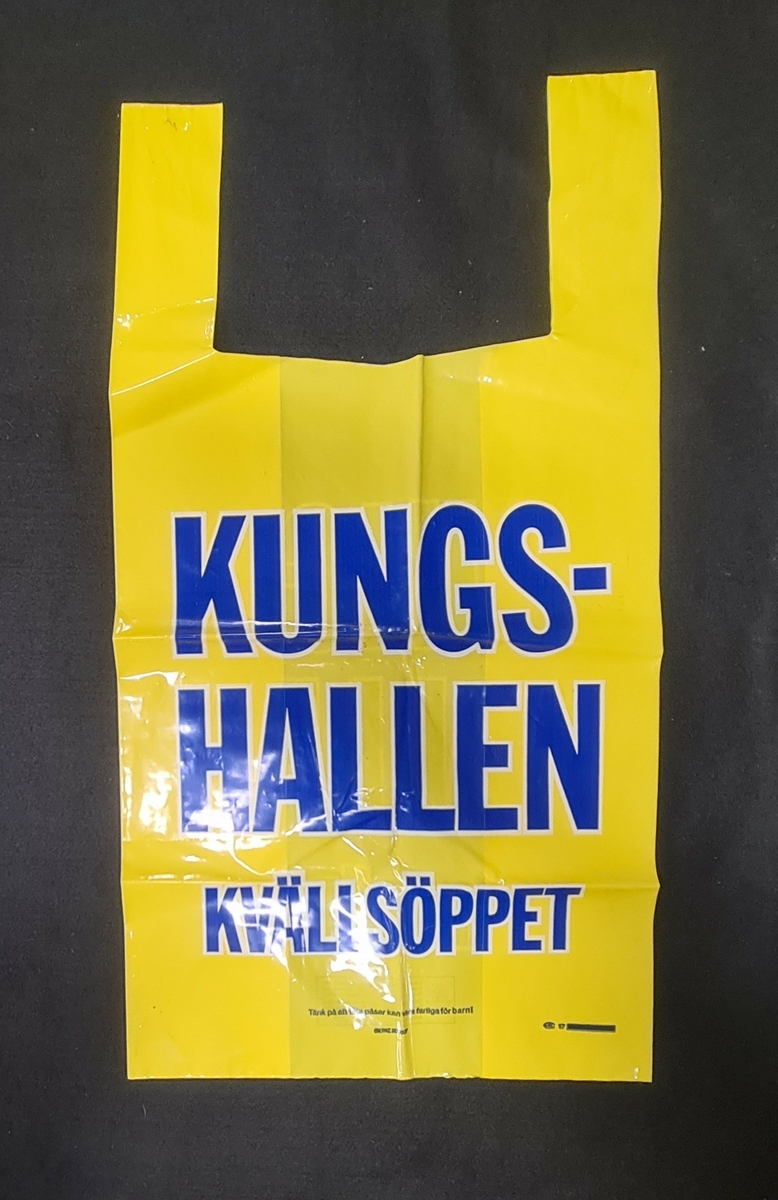 Plastkasse, gul, med logga från Kungshallen i Trollhättan. Text i blått. 

På kassen står:
Kungshallen
Kvällsöppet

Tänk på att påsar kan vara farliga för barn!
