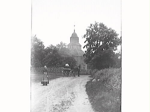En hästskjuts är på väg mot Okome gamla kyrka och i förgrunden står en kvinna med randigt förkläde vänd mot fotografen. Okome nya kyrka uppfördes 1891.