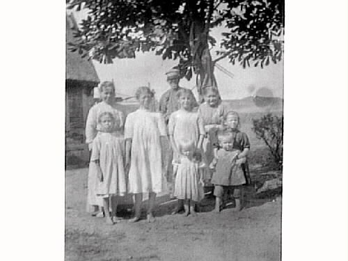 Ett äldre par med sju barn i olika åldrar under ett träd vid knuten av en byggnad i Ugglarp.