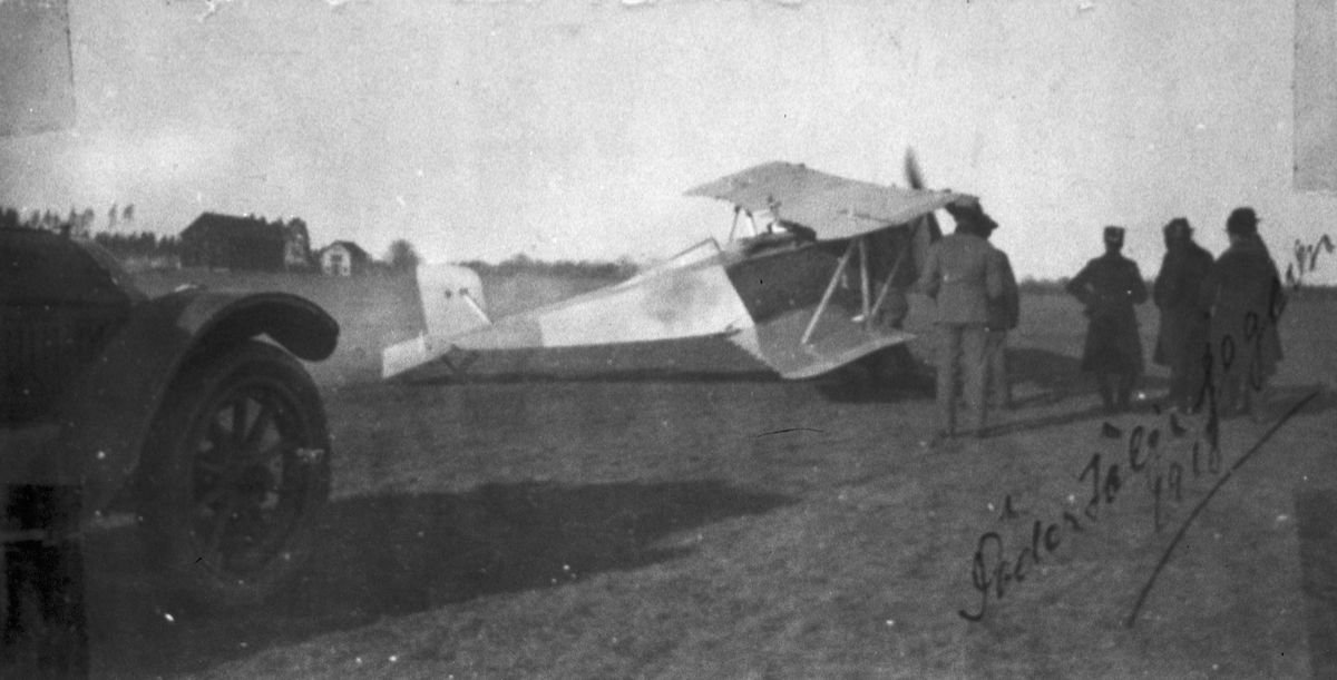Flygplan SW 15 "Lotterijagaren" på ett flygfält, 1918. Militärer runt planet. Vy snett bakifrån. I förgrunden syns en bil.