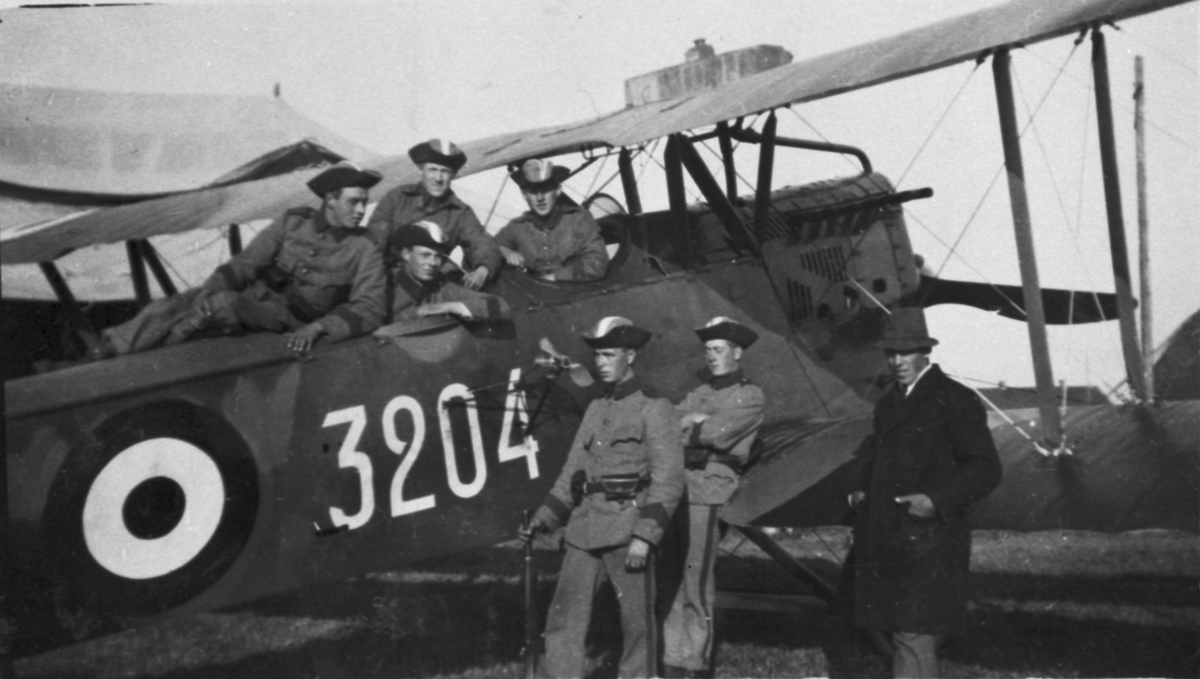 Sex militärer och en civilklädd man står vid ett övningsmärkt flygplan FVM S 21HL nummer 3204 under övning i Uppsala, 1926.
Vid fyran sitter ett propelleraggregat för drivande av filmseriekamera.