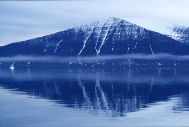 Ett fläckvis snötäckt berg i Sördalsbukta speglar sig i stilla vatten. En strimma av dis ligger strax ovanför vattenytan