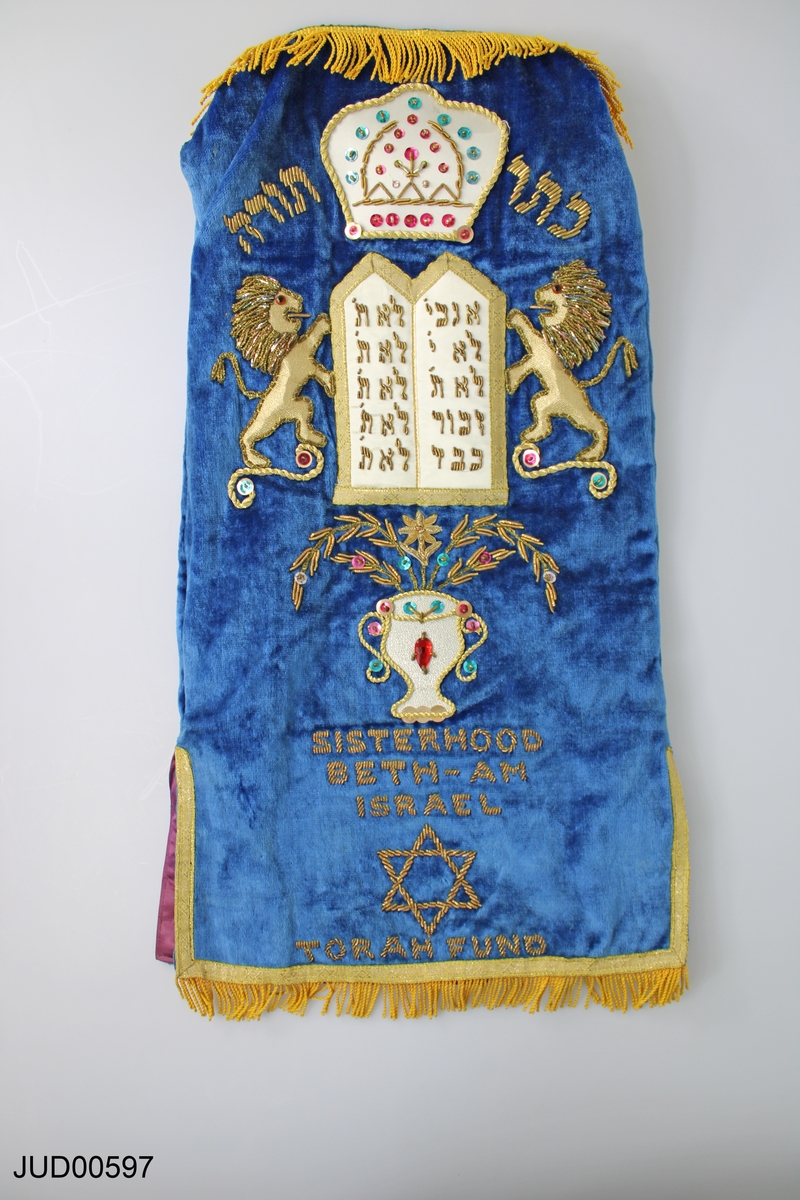 Torahrulle med tillhörande torahmantel. Torahmanteln tillverkad av blå sammet och dekorerad med guldbroderier i form av texten "Sisterhood Beth Israel" samt en vas med halvädelstenar. Själva torahrullen är tillverkad av pergament.