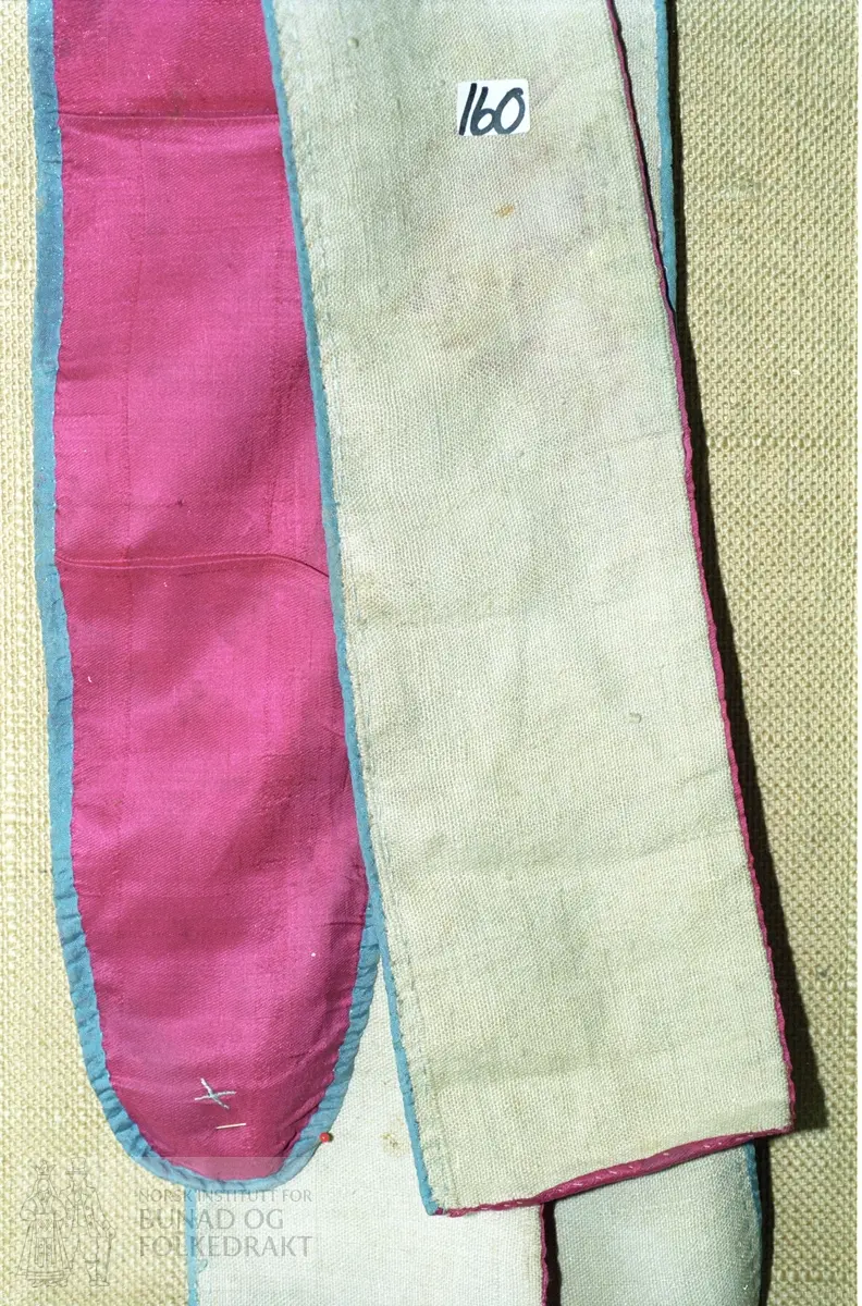 Raud silke kanta på eine langsida og delar av andre med blå silke. Fôra med linlerret. Avrunda i eine enden. Breidde:  11 - 12 cm, lengde:  ca. 280 cm.