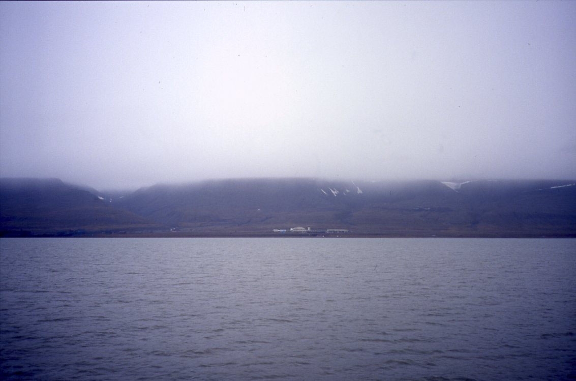 En grå dag över Isfjorden, låga moln över kustlinjens berg. I fjärran anas Longyearbyens flygplats.
