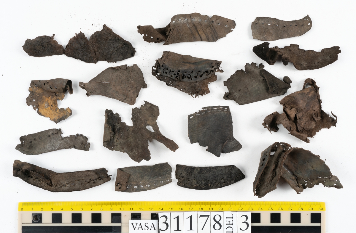 Runt 400 delar och fragment från skor i form av sulor, inlägg, ovanläder, klack, sidostycken, bakkappor, träpliggar, tråd mm. Troligtvis rör det sig om delar och fragment från flera skor och/eller stövlar. Delar av lädret har förstärkts med lim av konserveringsenheten. Merparten av lädret är spaltat samt har hål efter träpligg eller stygn.