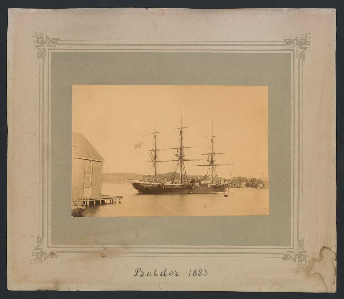 Bilden föreställer ångkorvetten Balder som ligger till ankars i Arendal. Fartygets syns från babordssidan med bärgade segel. Vid förskeppet ligger flera skeppsbåtar från aktern hänger det ner flera repstege.
