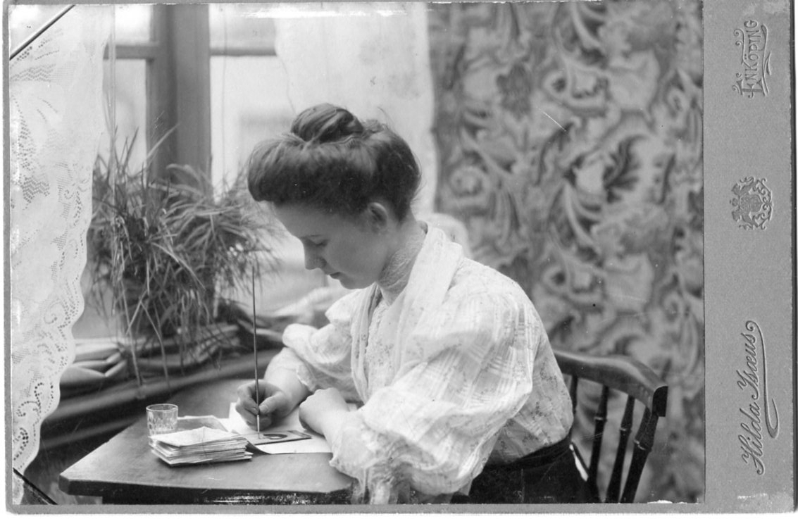 Kabinettsfotografi: en ung kvinna sitter vid ett skrivbord med en tunn pensel i handen. Hon retuscherar kabinettsfotografier.