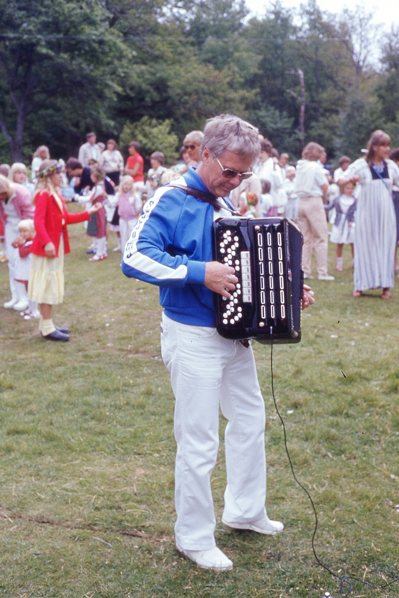 Barn och vuxna dansar kring stången vid midsommarfirande på Ekensås cirka 1982. Spelmannen Frank Efraimsson spelar dragspel.