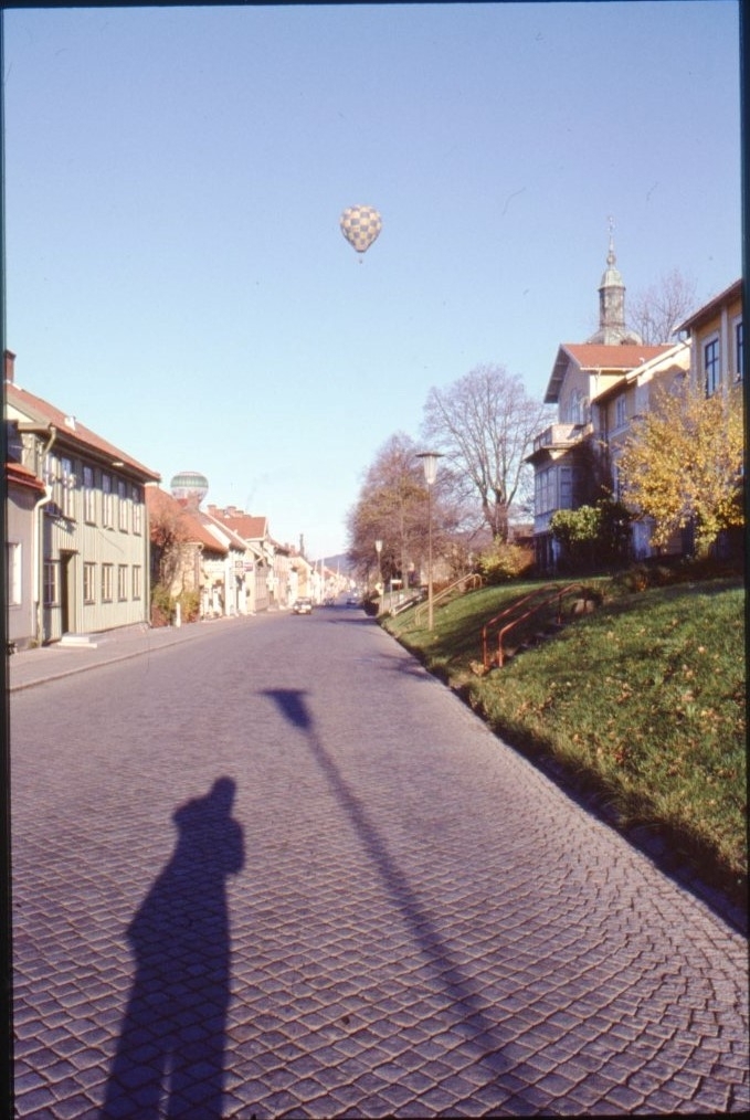 En grön-vit rutig ballong i luften. Fotograferad från Brahegatans norra del mot söder. Fotografens skugga syns i gatan.