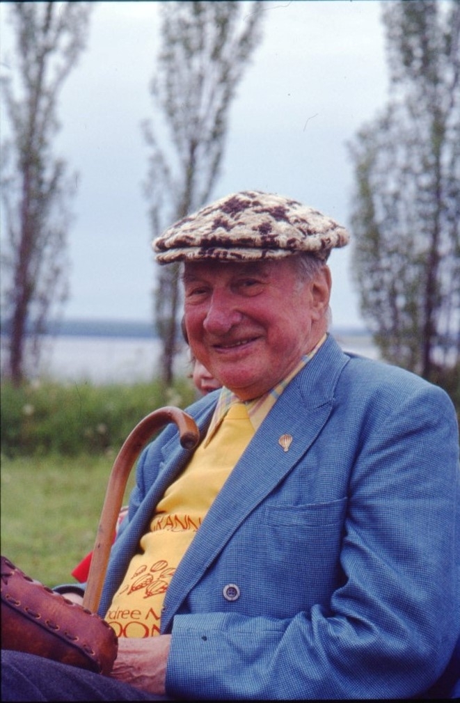 Porträtt av en äldre skrattande man. Här keps, gul tröja med ballonger på och blå kavaj, på kavajslaget sitter en ballong-pin. Han håller i en käpp.