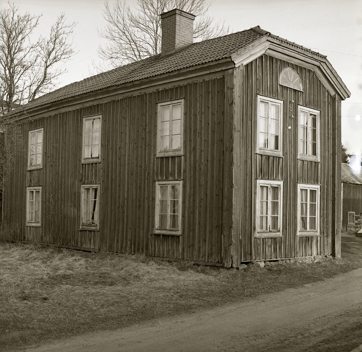 Baksidan av en ganska förfallen gammal hälsingegård, Söderala 1973. Timmerhus klätt med träpanel. Valmat tak med tegel. Fönster med spröjs och sex rutor och längst upp ett halvmånefönster. Vid gaveln går en smal grusväg.
