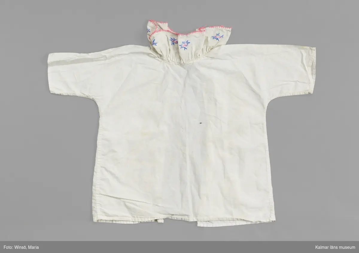 KLM 46489:16. Skjorta, barnskjorta, textil. Barnskjorta av vitt bomullstyg med öppning i ryggen. Knyts med vitt bomullsband i ryggen. Volangkrage med broderade små stjärnliknande mönster i rosa och blått. Broderade stygn runt kragens kant i rosa.