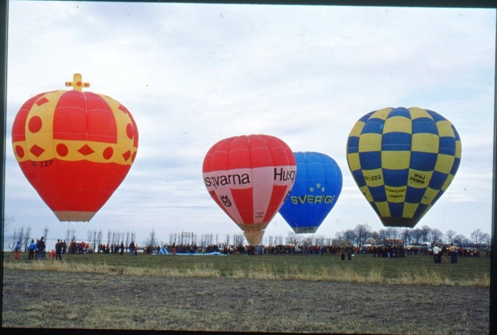 Fyra färgglada luftballonger på gräsplanen nere vid Vättern i Gränna. Den vänstra, i färger som en krona, är Riksäpplet från Uppsala. Därefter den rödvita "Örnen II Gränna märkt "Husqvarna", en blå märkt "Sverige" samt en blågul-rutig märkt "Spola kröken". De senare tillhör Ballongklubben Andrée i Gränna, Skänninge Ballongklubb samt Tekniska museet. En folksamlig runt ballongkorgarna och i bakgrunden.