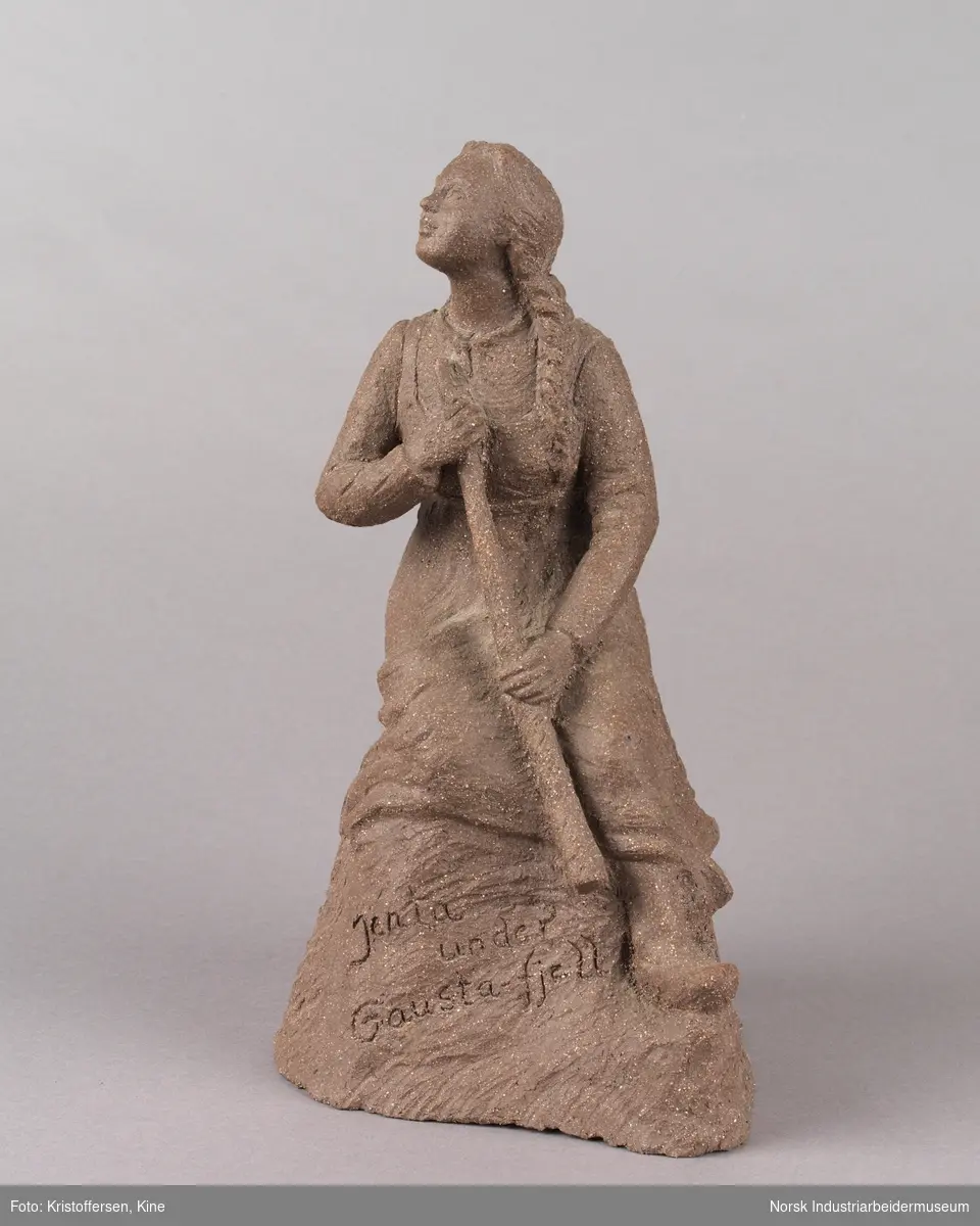 Skulptur laget av leire. Figur, en kvinne med fletter og iført en kjole sitter på en stein med en lur i hendene. Kvinnen vender blikket oppover. Skulpuren er hul.