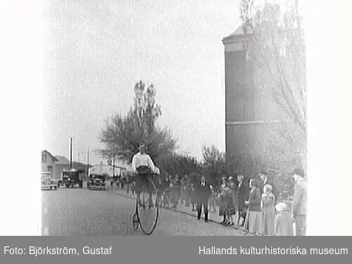 Uppvisning där en man cyklar på en gammal höghjuling, sannolikt på Cykelns Dag i Varberg. Här rullar han förbi åskådrna på Södra vägen vid Rosenfredsskolan. Enligt fotografens liggare är fotot från Sexdagarsloppet det året. Det var en årlig amatörcykeltävling som startades 1924.